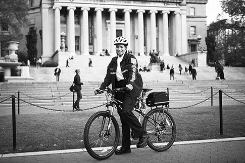 A female public safety officer straddles a bicycle on College Walk with Low Library in the background on a sunny day